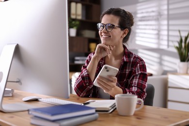 Happy woman holding smartphone and working with computer at desk indoors. Home office