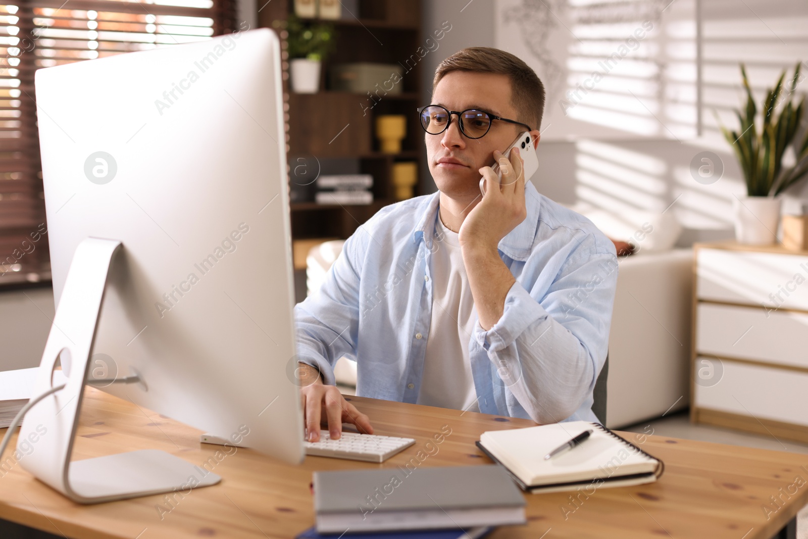 Photo of Handsome man talking by smartphone at desk with computer indoors. Home office