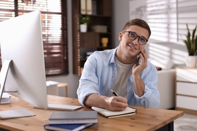 Happy man talking by smartphone at desk with computer indoors. Home office