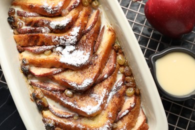 Photo of Freshly baked bread pudding in baking dish and custard cream on dark table, top view