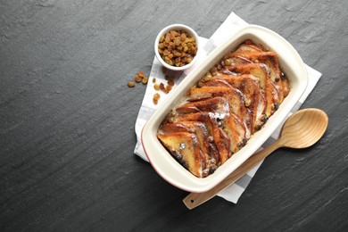 Photo of Freshly baked bread pudding in baking dish, raisins and wooden spoon on dark textured table, top view. Space for text