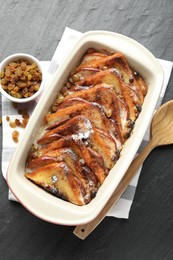 Photo of Freshly baked bread pudding in baking dish, raisins and wooden spoon on dark textured table, top view