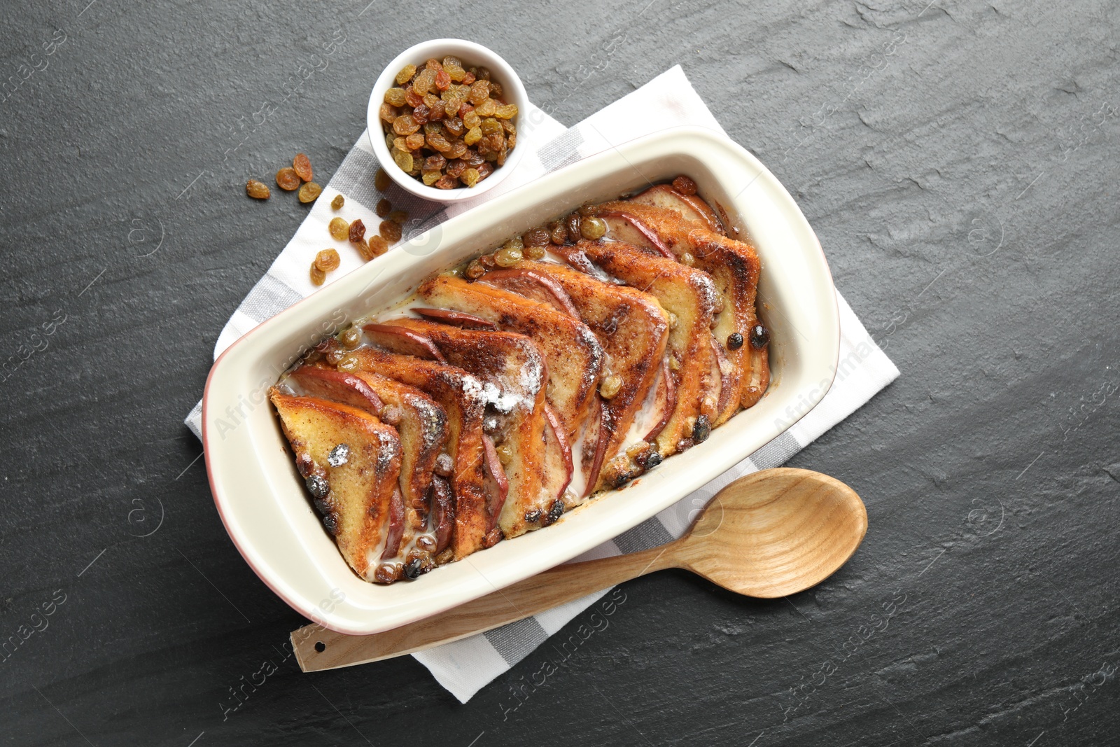 Photo of Freshly baked bread pudding in baking dish, raisins and wooden spoon on dark textured table, top view