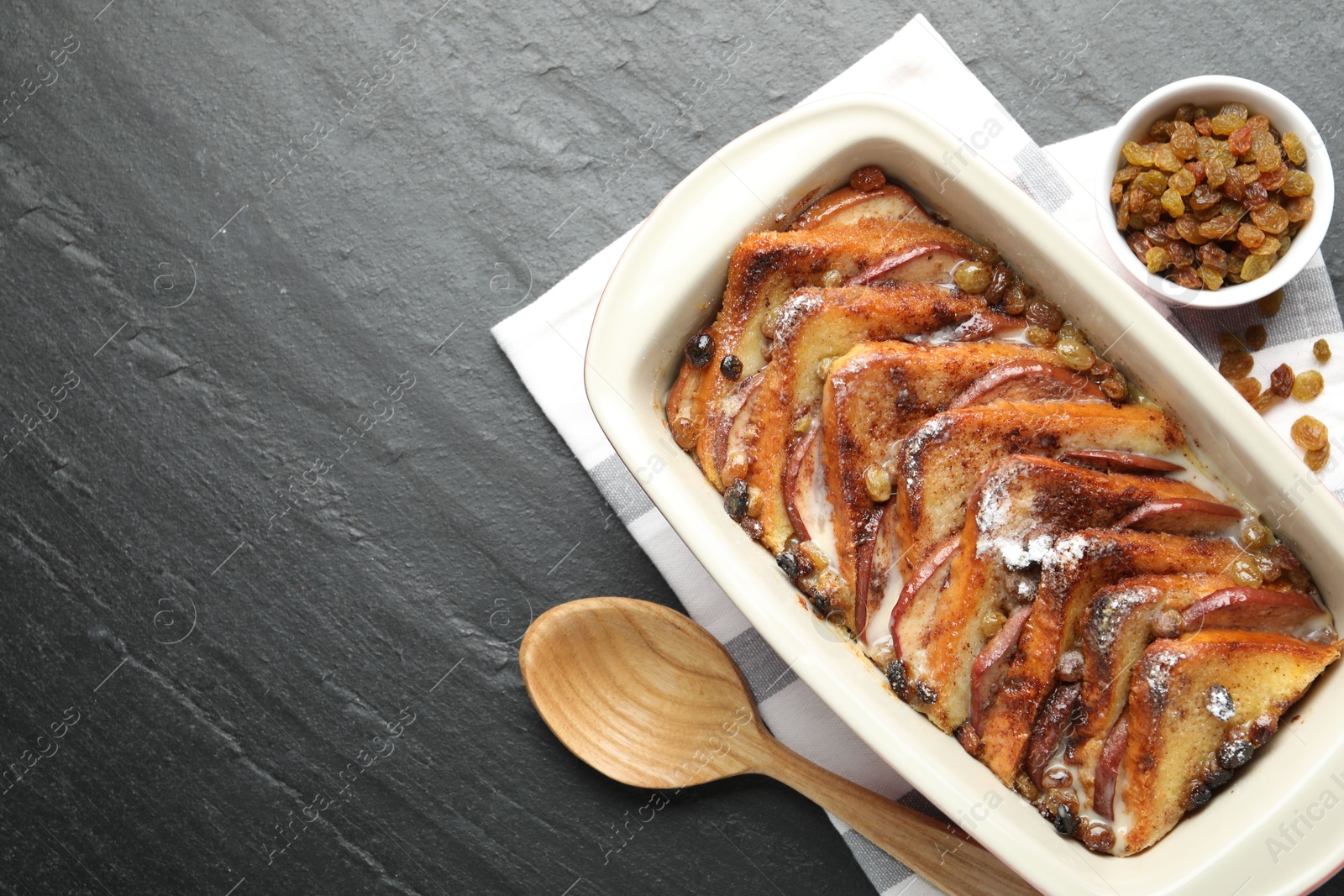 Photo of Freshly baked bread pudding in baking dish, raisins and wooden spoon on dark textured table, top view. Space for text