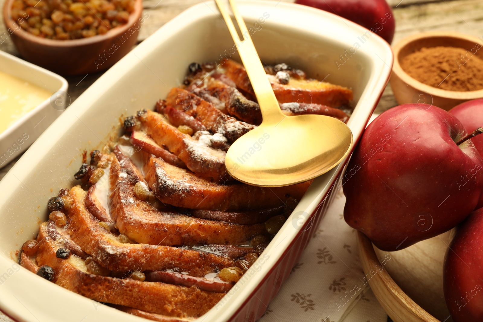 Photo of Freshly baked bread pudding in baking dish, spoon and apples on table, closeup