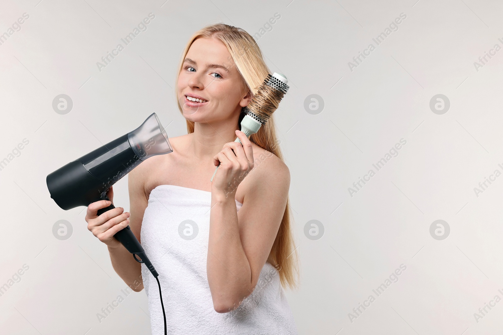 Photo of Beautiful young woman styling her hair with hairdryer and brush on light grey background