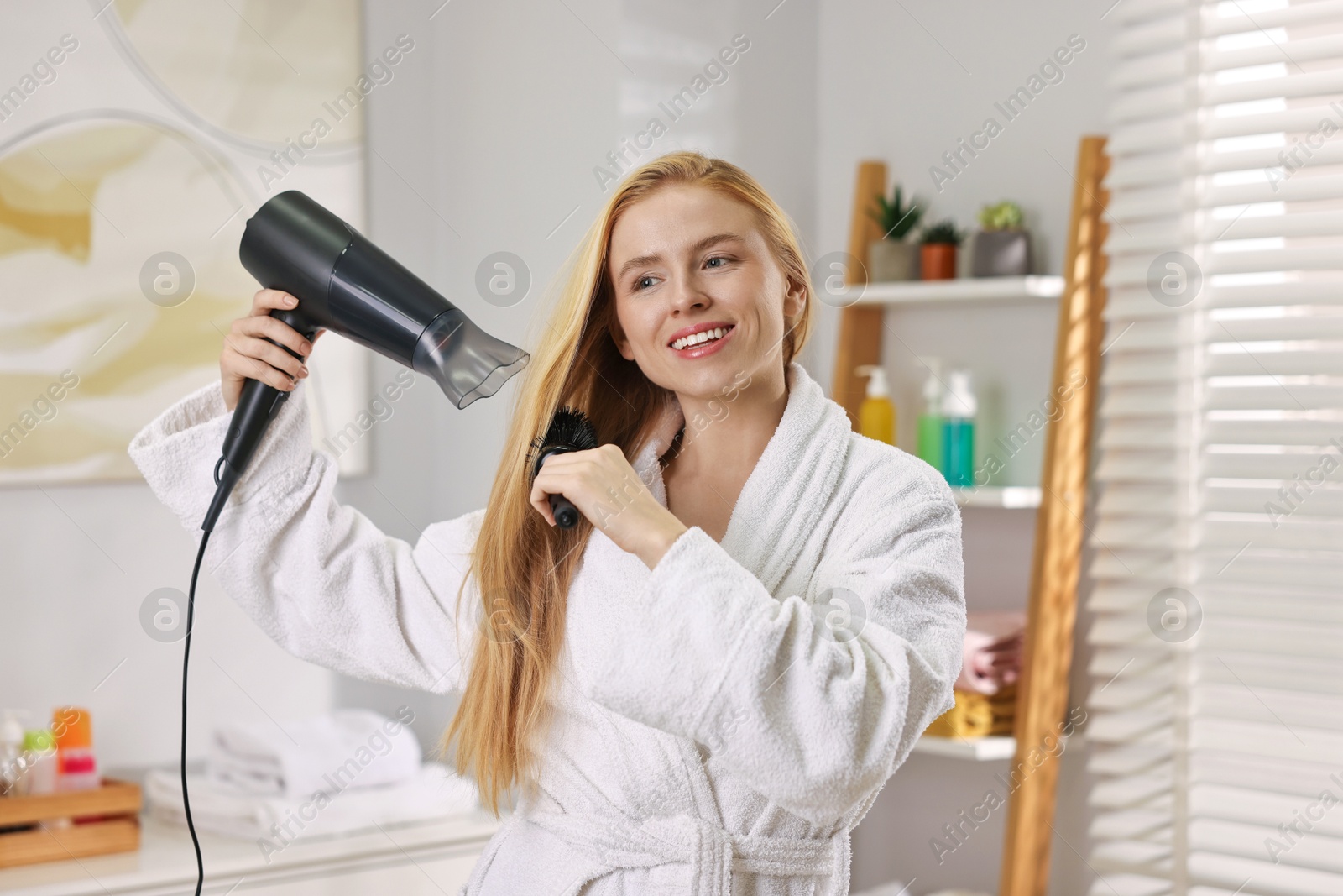 Photo of Beautiful young woman styling her hair with hairdryer and brush in bathroom