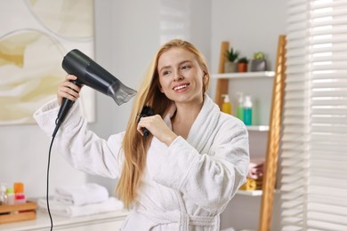 Beautiful young woman styling her hair with hairdryer and brush in bathroom