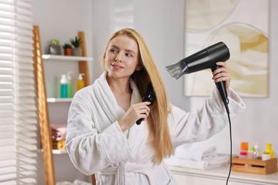 Beautiful young woman styling her hair with hairdryer and brush in bathroom