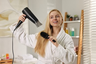 Beautiful young woman styling her hair with hairdryer and brush in bathroom