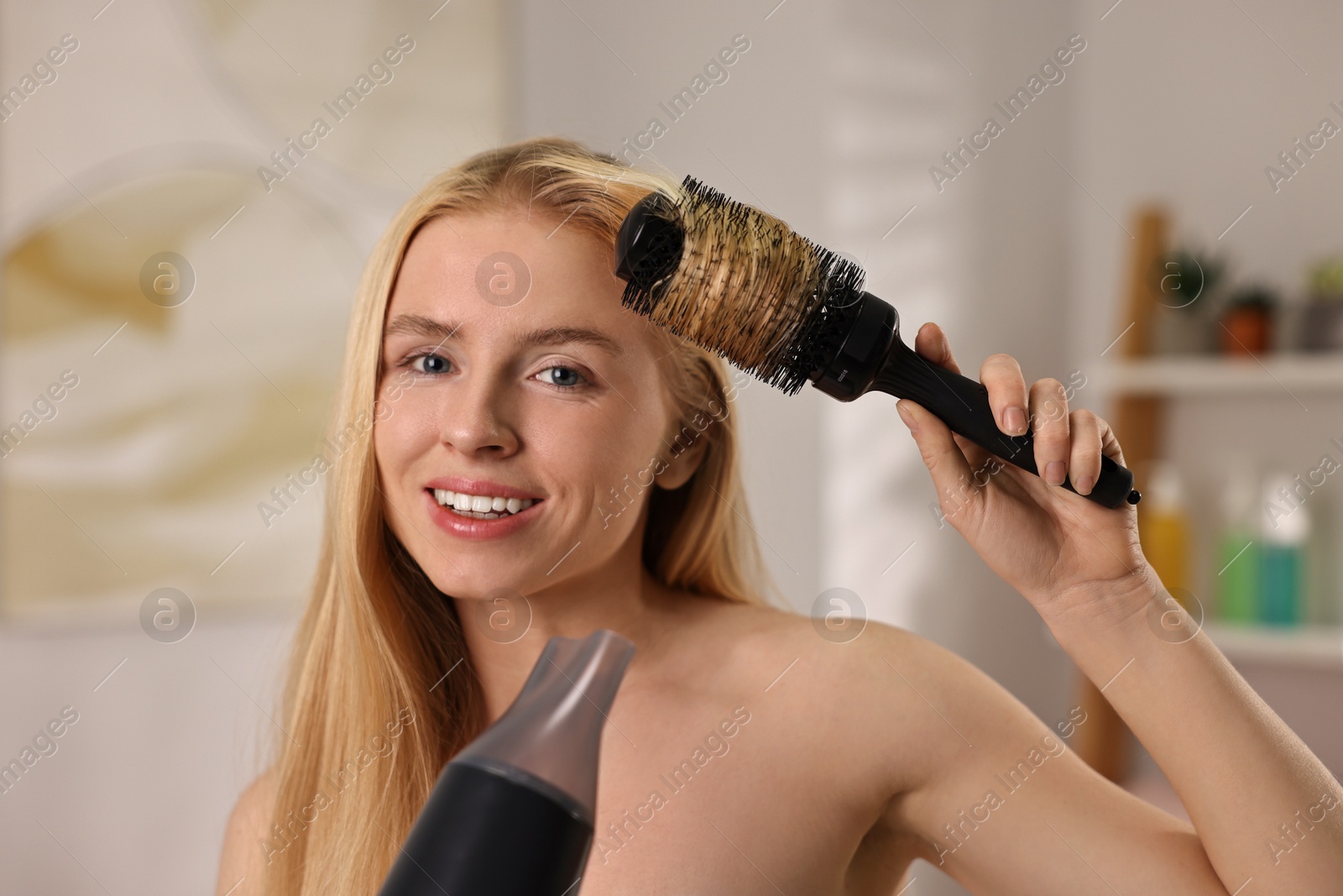 Photo of Beautiful young woman styling her hair with hairdryer and brush in bathroom