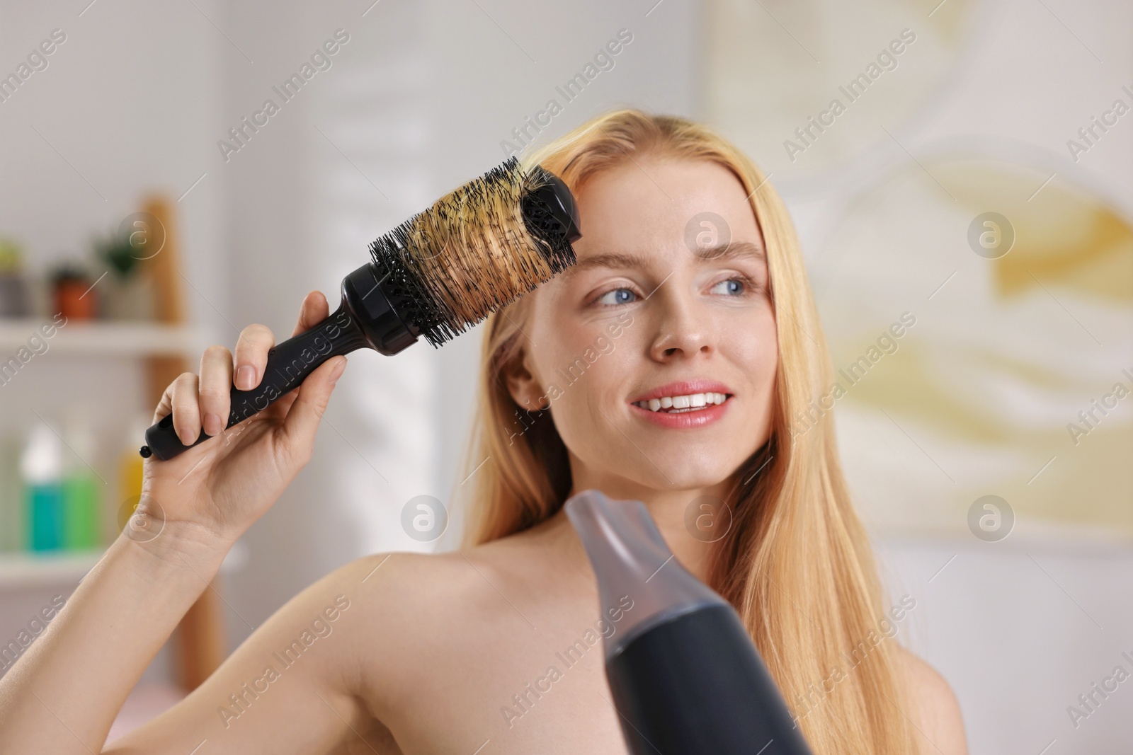 Photo of Beautiful young woman styling her hair with hairdryer and brush in bathroom