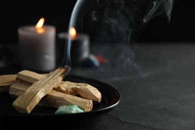 Photo of Smoldering palo santo stick, gemstone and burning candles on black table, closeup. Space for text