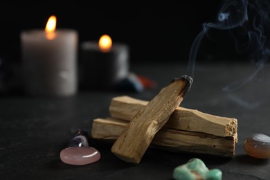 Photo of Smoldering palo santo stick, gemstones and burning candles on black table, closeup