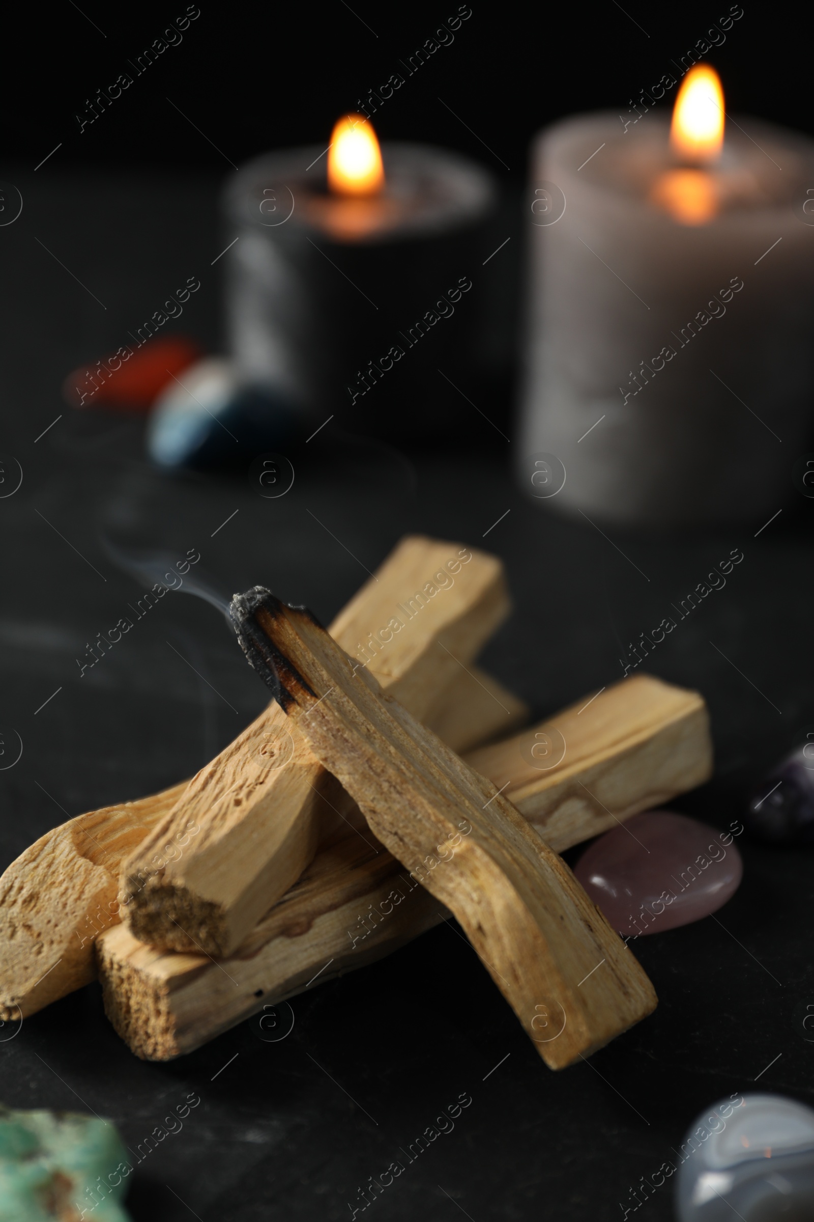 Photo of Smoldering palo santo stick, gemstones and burning candles on black table, closeup