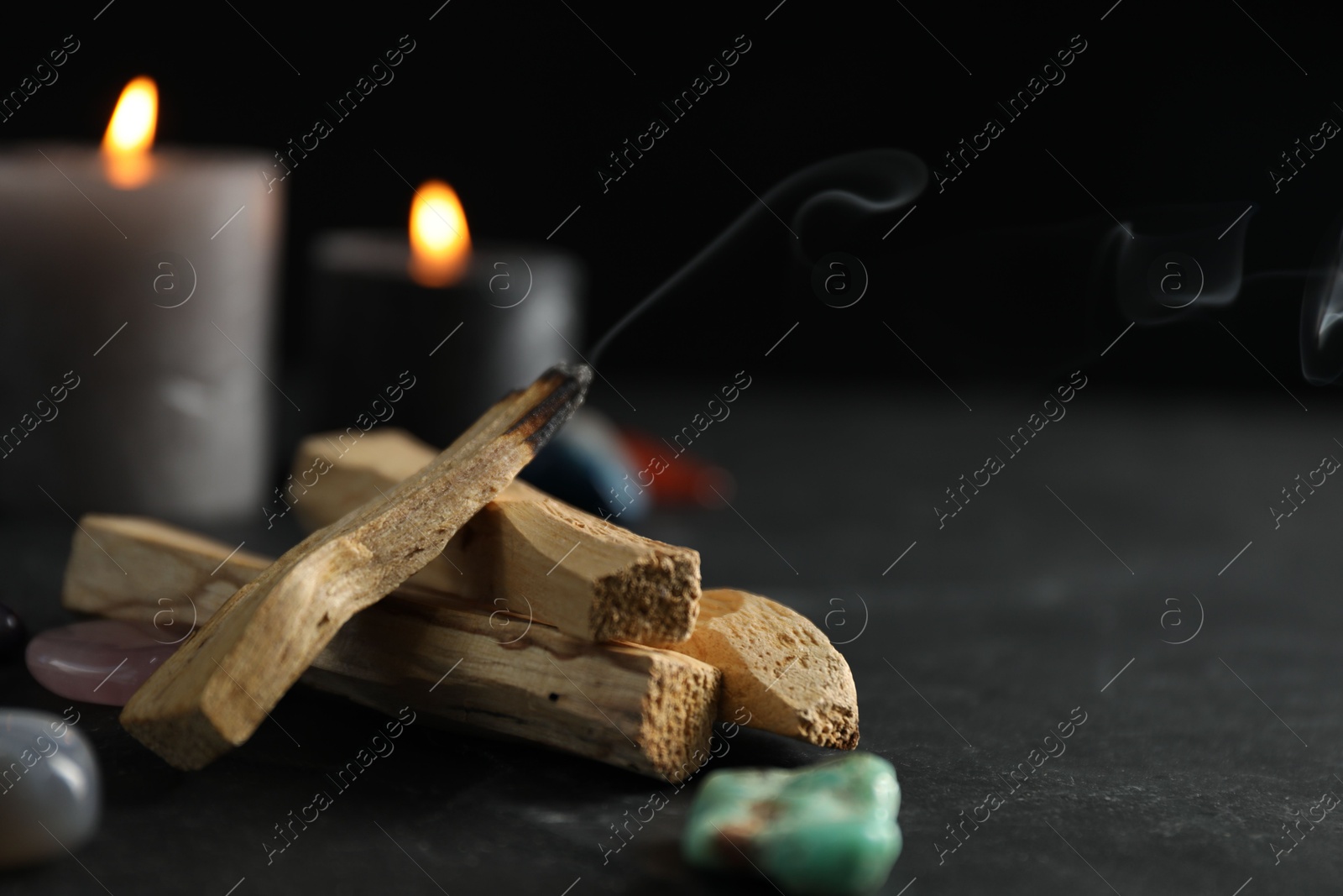 Photo of Smoldering palo santo stick, gemstones and burning candles on black table, closeup. Space for text