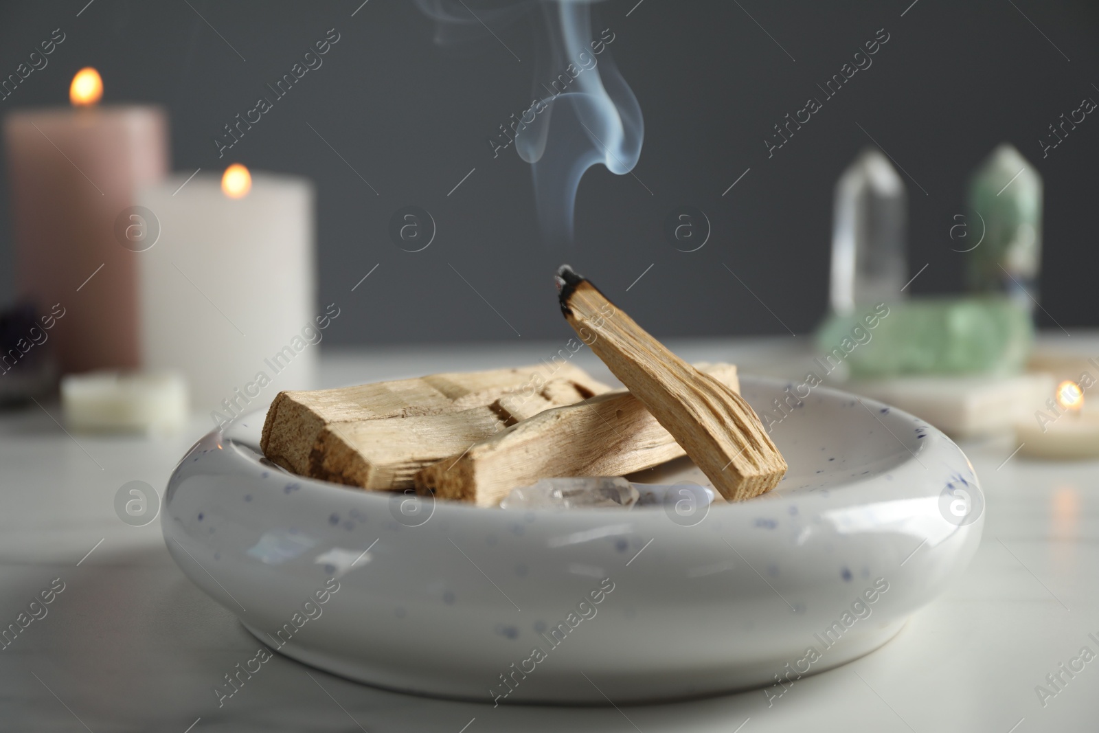 Photo of Smoldering palo santo stick, gemstones and burning candles on white table, closeup