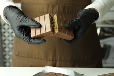 Photo of Woman breaking tasty Dubai chocolate with pistachios and knafeh at light table indoors, closeup