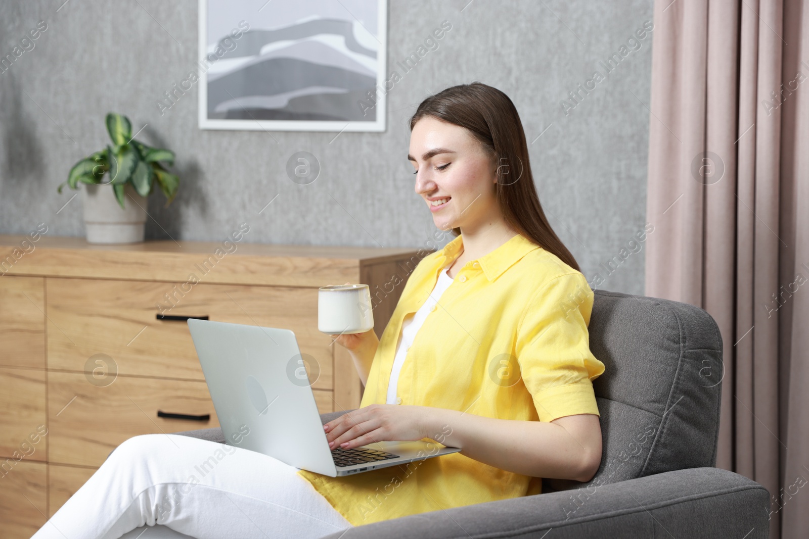 Photo of Young woman with cup of drink working on laptop at home