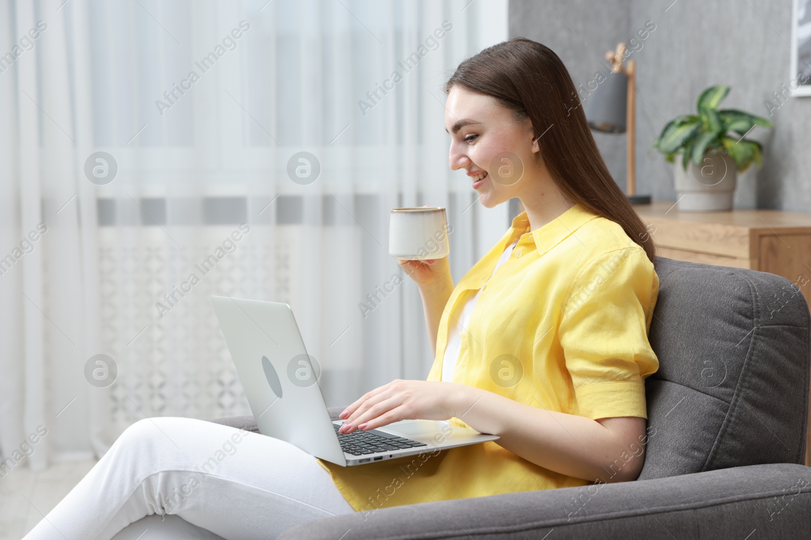 Photo of Young woman with cup of drink working on laptop at home