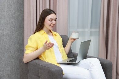 Photo of Happy woman with cup of drink having video chat via laptop in armchair at home