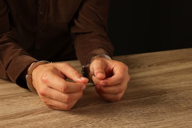 Photo of Man in metal handcuffs at wooden table against black background, closeup