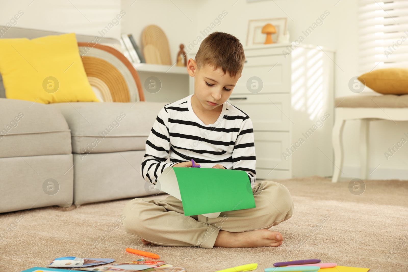 Photo of Creating vision board. Little boy cutting green paper with scissors on floor at home