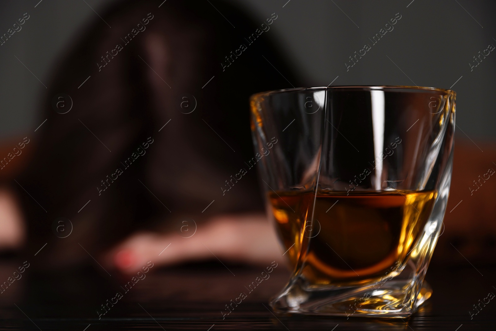 Photo of Alcohol addiction. Woman at wooden table indoors, focus on glass of whiskey