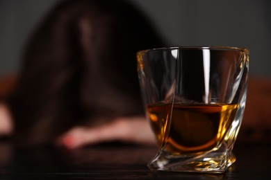 Photo of Alcohol addiction. Woman at wooden table indoors, focus on glass of whiskey