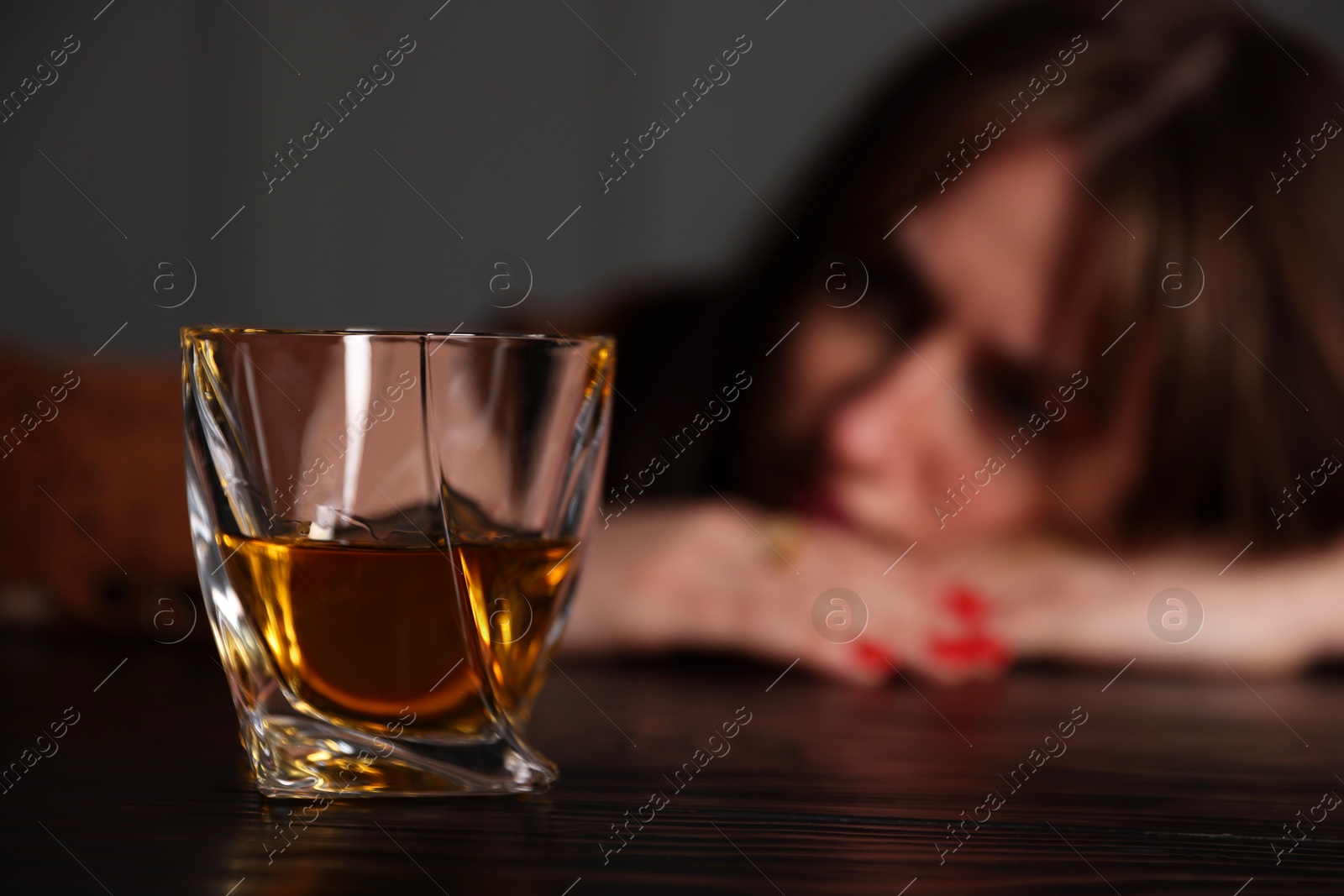 Photo of Alcohol addiction. Woman at wooden table indoors, focus on glass of whiskey