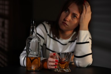 Photo of Alcohol addiction. Woman with glass of whiskey and bottle at table indoors, selective focus