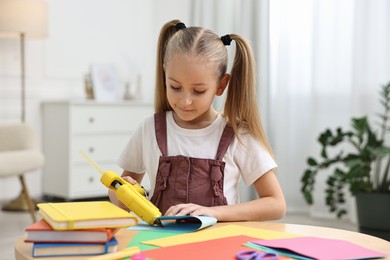 Photo of Little girl with hot glue gun and color paper making craft at table indoors