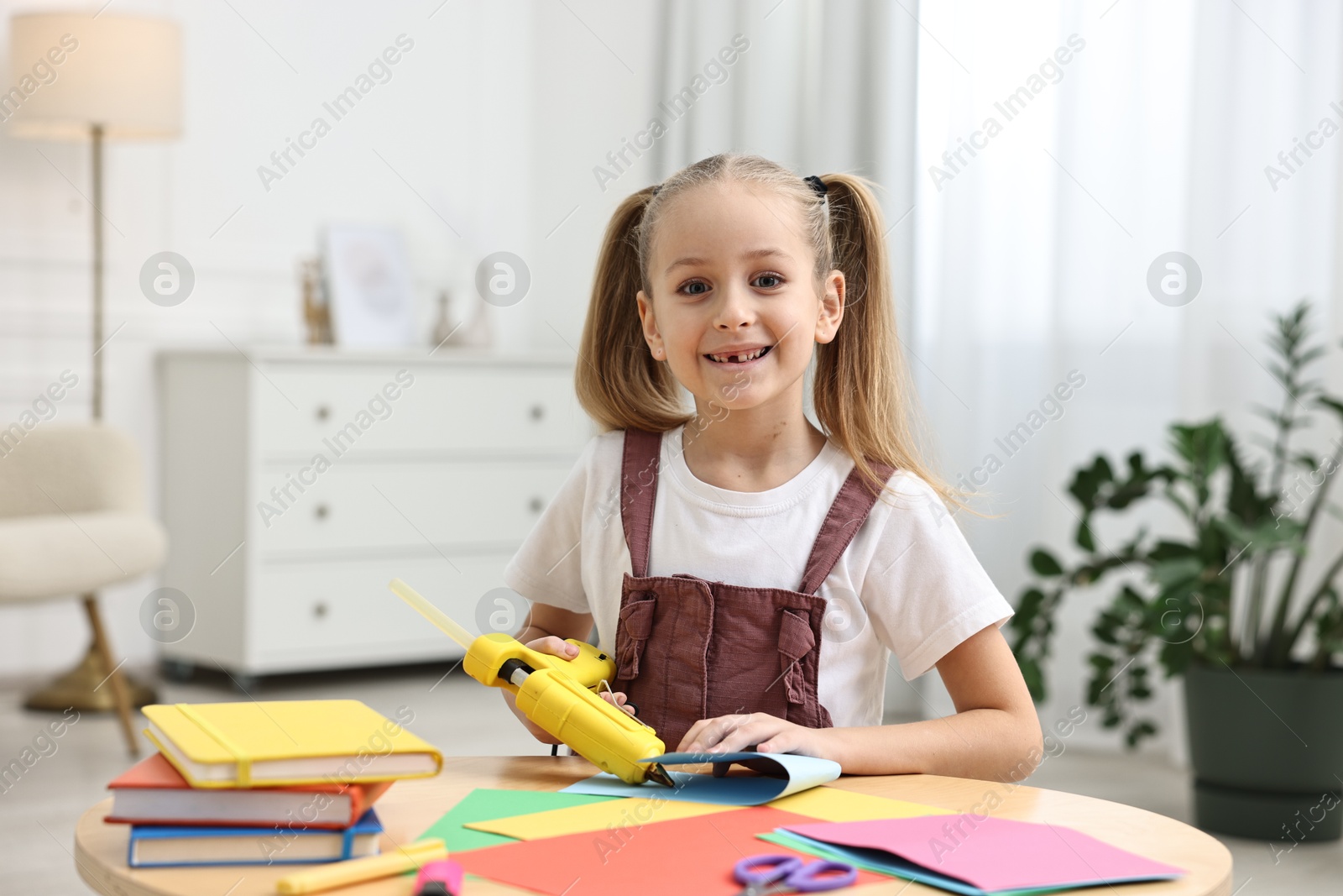 Photo of Smiling girl with hot glue gun and color paper making craft at table indoors