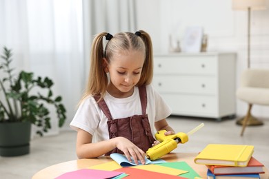 Photo of Little girl with hot glue gun and color paper making craft at table indoors