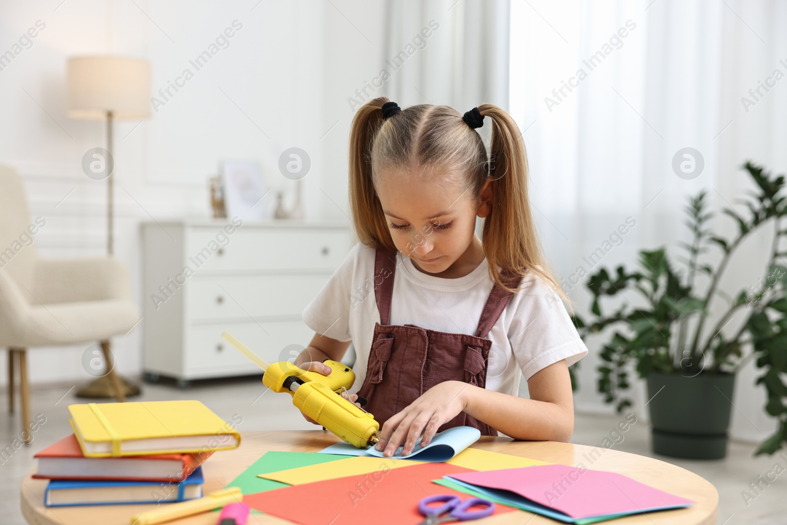 Photo of Little girl with hot glue gun and color paper making craft at table indoors