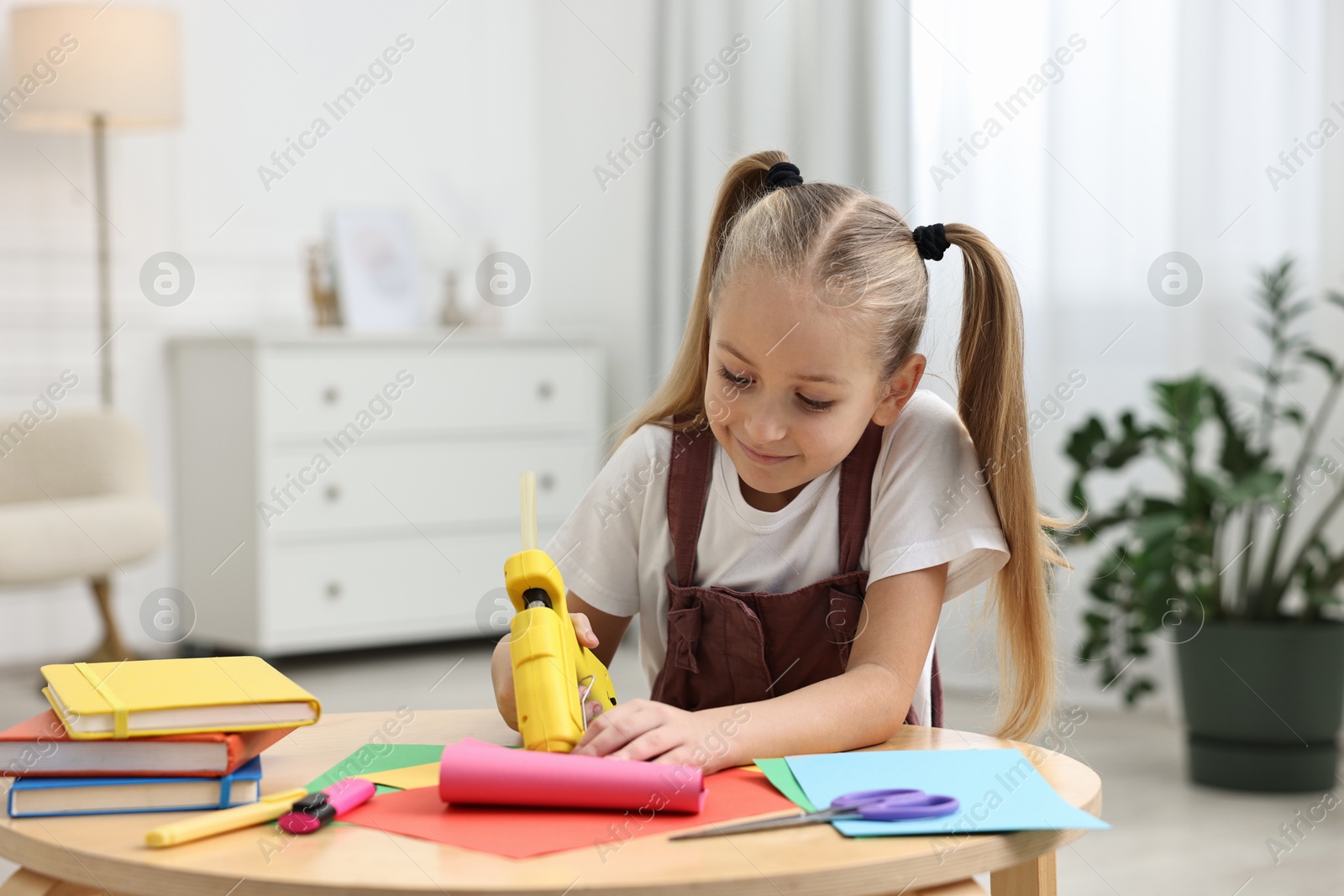 Photo of Little girl with hot glue gun and color paper making craft at table indoors