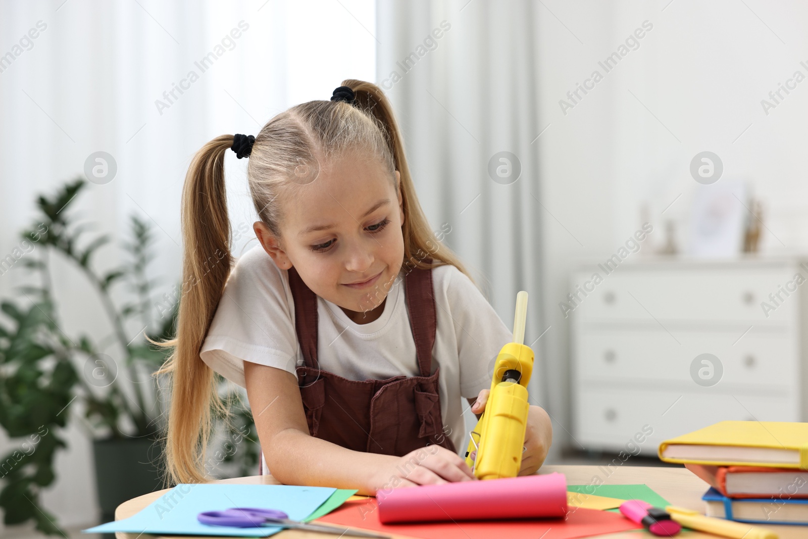 Photo of Little girl with hot glue gun and color paper making craft at table indoors