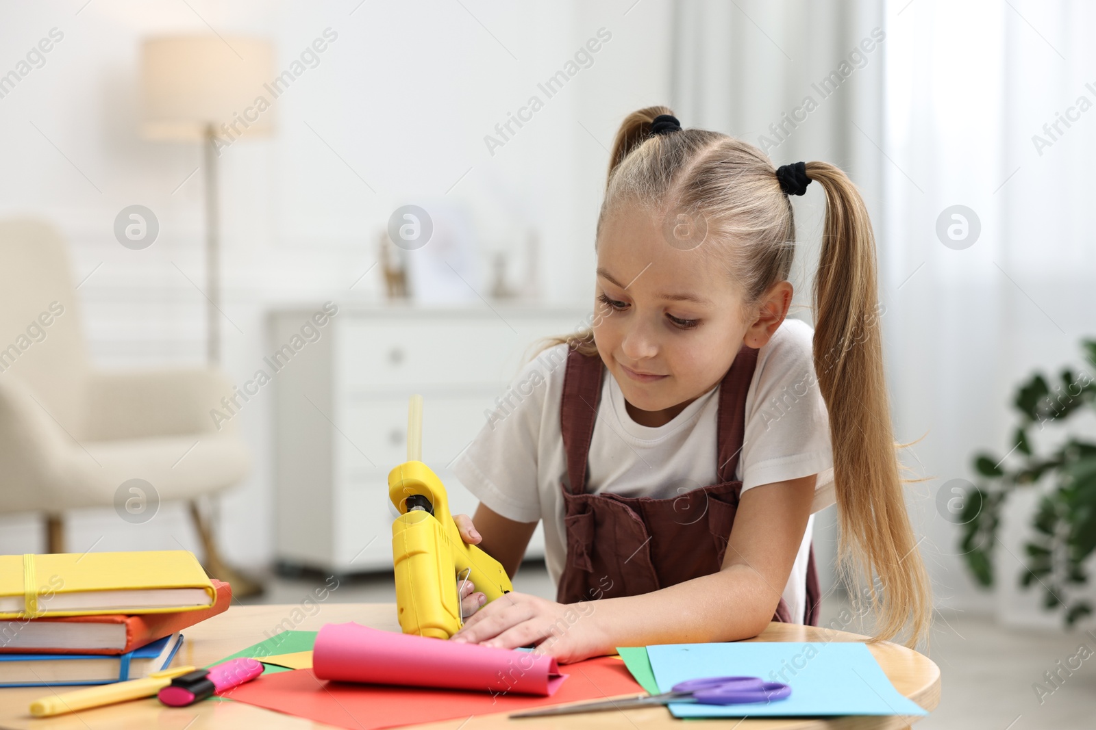 Photo of Little girl with hot glue gun and color paper making craft at table indoors