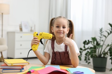 Photo of Little girl with hot glue gun and color paper showing thumbs up at table indoors