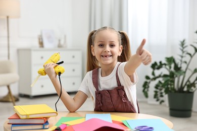 Photo of Smiling girl with hot glue gun and color paper showing thumbs up at table indoors