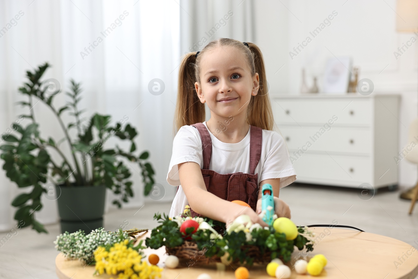 Photo of Little girl with hot glue gun creating Easter composition at table indoors