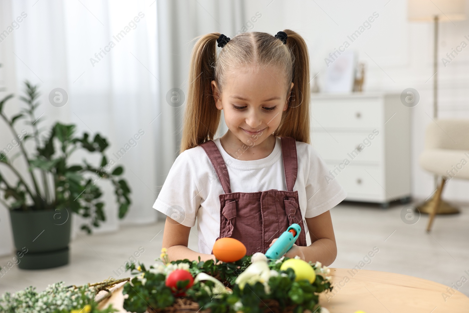 Photo of Little girl with hot glue gun creating Easter composition at table indoors