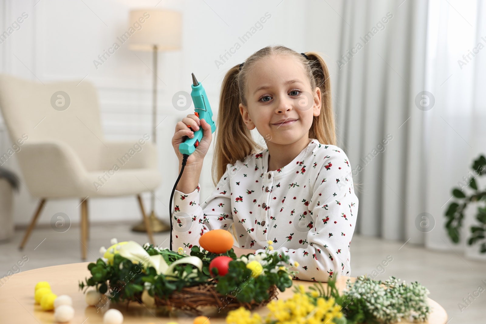 Photo of Little girl with hot glue gun creating Easter composition at table indoors