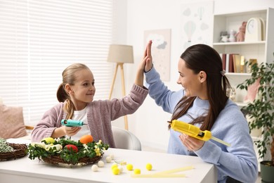 Mother with glue gun giving high five to daughter at table indoors