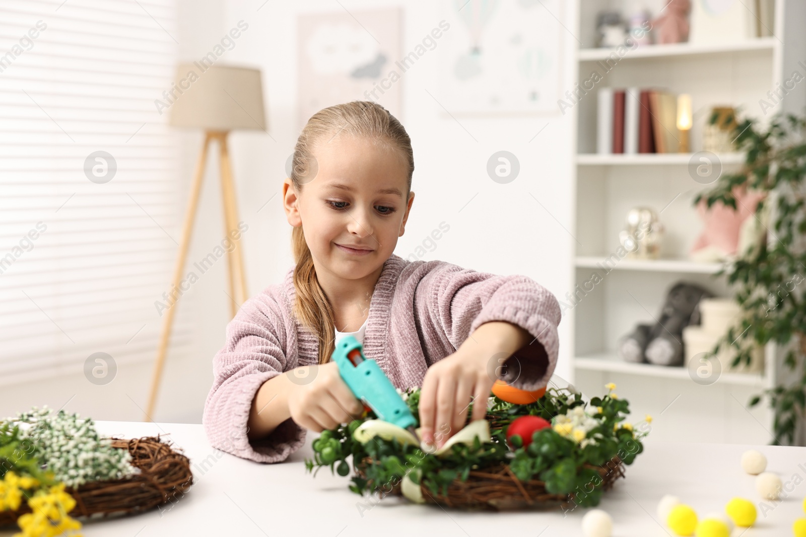 Photo of Little girl with hot glue gun creating Easter composition at table indoors