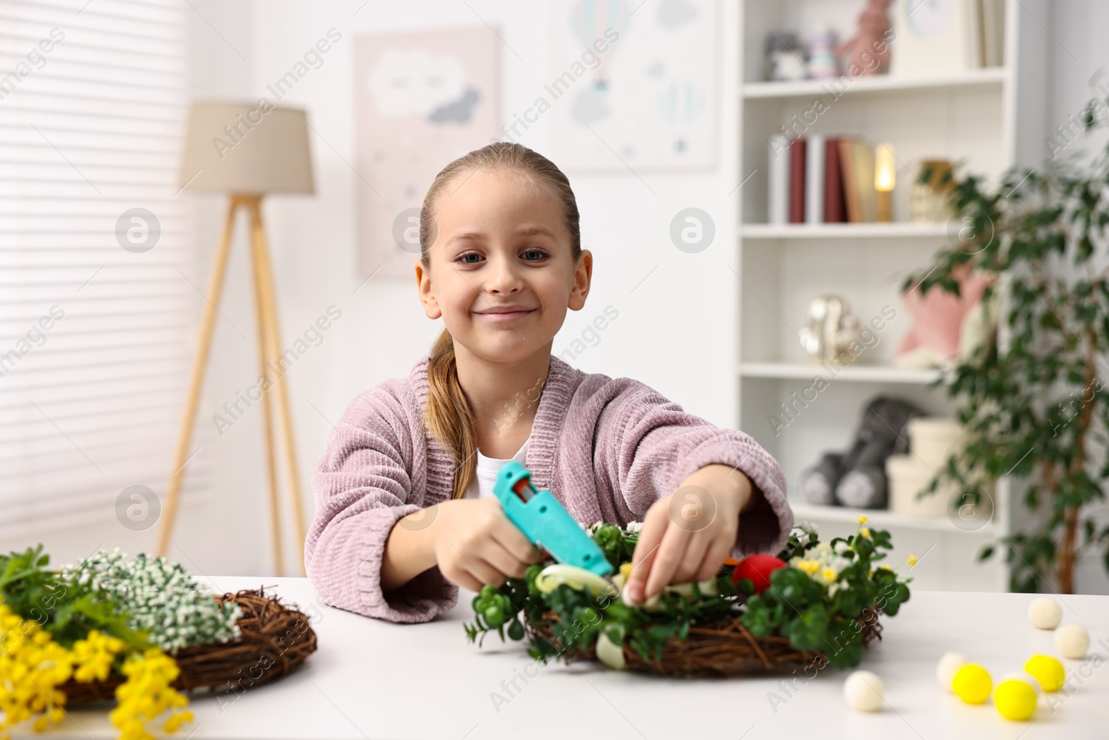 Photo of Little girl with hot glue gun creating Easter composition at table indoors