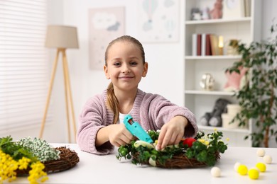 Photo of Little girl with hot glue gun creating Easter composition at table indoors