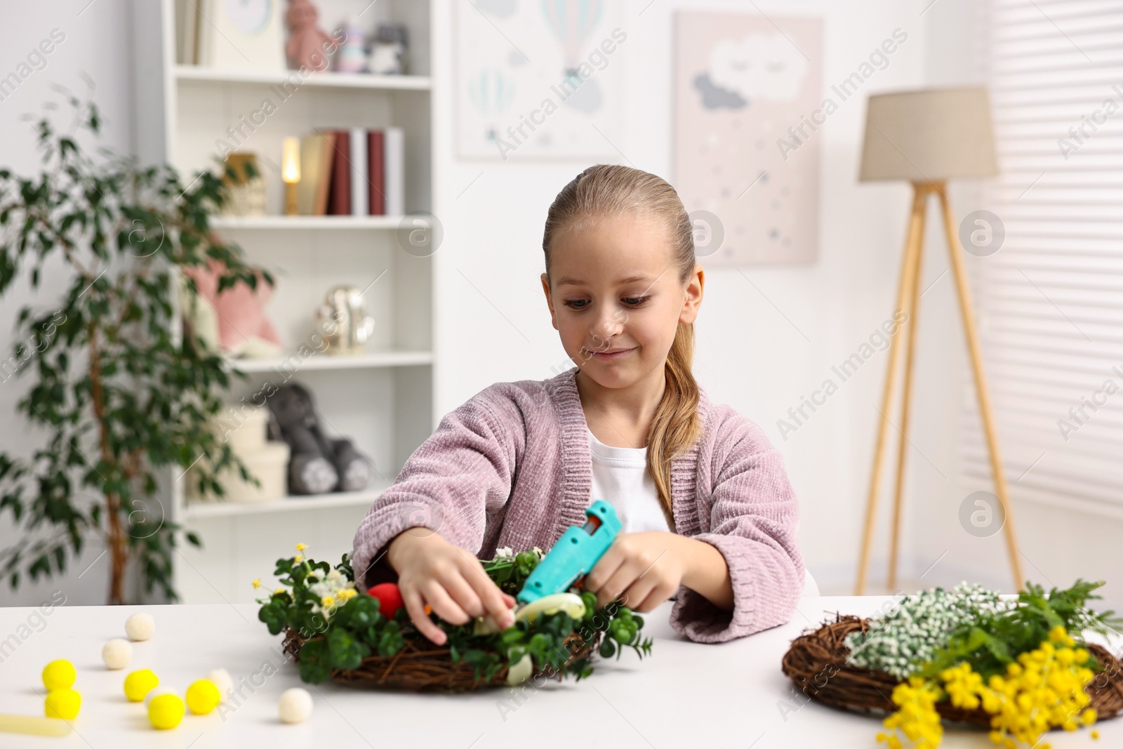 Photo of Little girl with hot glue gun creating Easter composition at table indoors