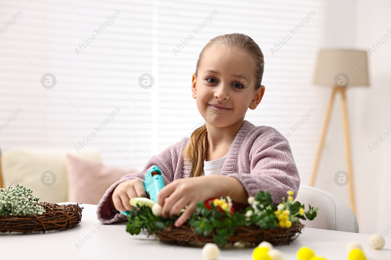 Photo of Little girl with hot glue gun creating Easter composition at table indoors