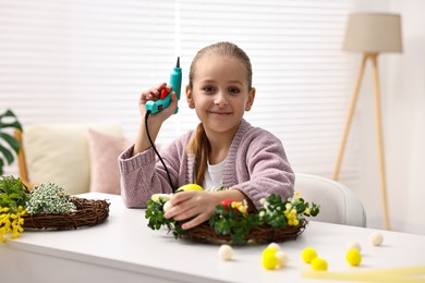 Photo of Little girl with hot glue gun creating Easter composition at table indoors